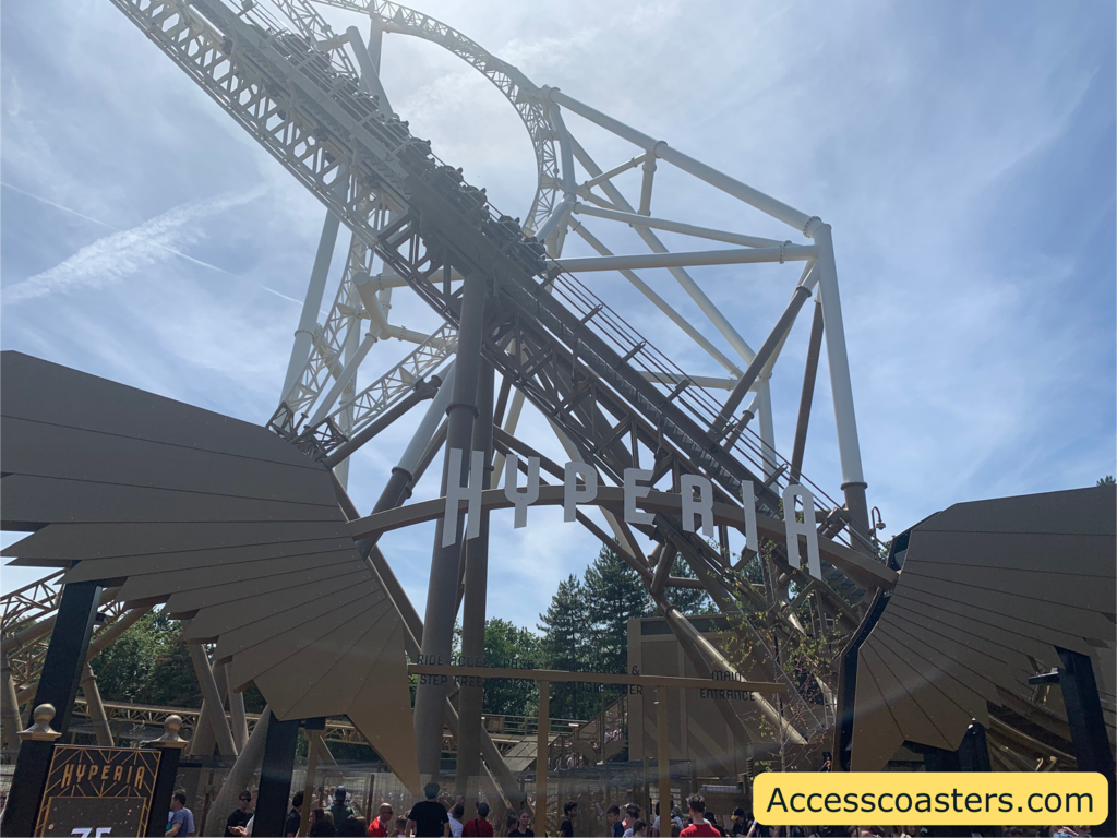 Image of Hyperia queue line entrance. in the image you can see the Hyperia entrance sign with gold wings eather side. you can also see the track above with a train going around.