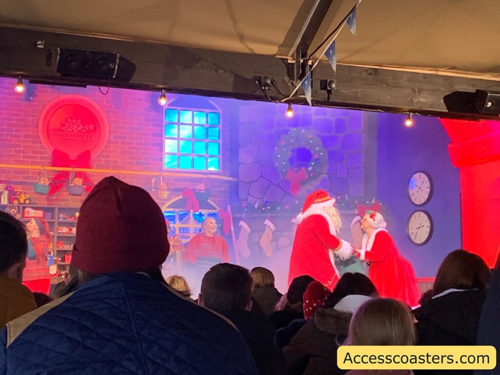 The image shows a festive stage performance with a Christmas theme. In the center, Santa Claus and Mrs. Claus are interacting with each other. They are both dressed in traditional red and white outfits. The background is designed to look like a cozy room with a brick wall, a wreath, and stockings hanging by a stone fireplace. There are shelves with toys and decorations, and a "Spirit-O-Meter" on the wall. The audience is visible in the foreground, watching the performance. The atmosphere is warm and festive, with Christmas lights and decorations enhancing the scene. In the bottom right corner, there is a watermark that reads "Accesscoasters.com".