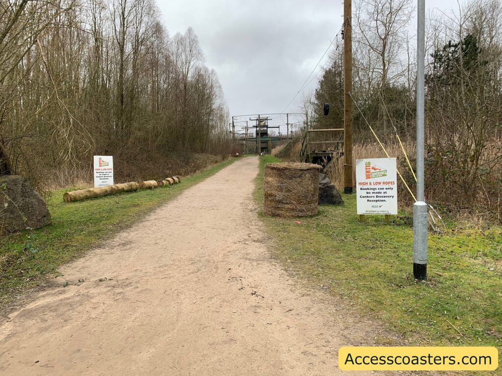 A dirt path runs through a wooded area with leafless trees on both sides. On the left side of the path, there are logs placed on the grass, and a sign that reads "Go Ape! HIGH & LOW ROPES Bookings can only be made at Conkers Discovery Reception." On the right side, there is another similar sign next to a pole, and a large coil of rope is placed on the grass. In the background, there is a structure that appears to be part of a ropes course. The sky is overcast, and the overall setting suggests an outdoor adventure or activity area. In the bottom right corner, there is a yellow rectangle with the text "Accesscoasters.com".