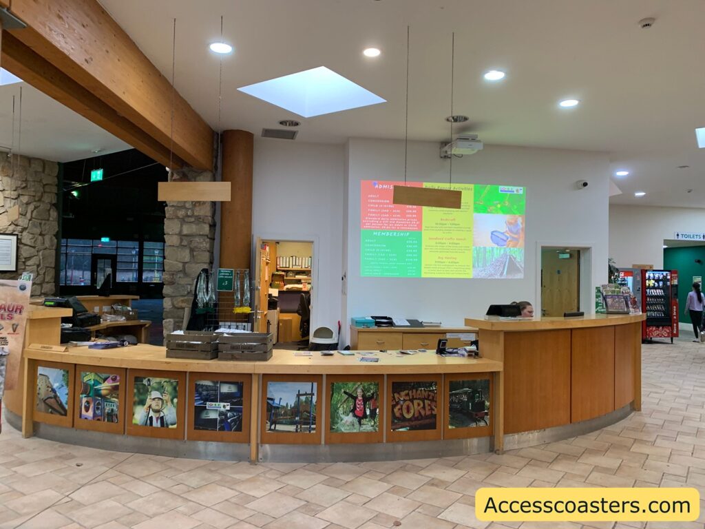 A reception area with a curved wooden desk. Behind the desk, a person is seated, and there are various items like brochures and a computer. On the wall behind the desk, a projector displays information about "Admission" and "Membership" with prices and activities like "Rockwall" and "Woodland Coffee Mornings." The wall also features a stone pillar and wooden beams. To the right, there is a vending machine and a sign indicating the direction to the toilets. The floor is tiled, and the ceiling has recessed lighting and skylights. The bottom of the image has a watermark that reads "Accesscoasters.com."