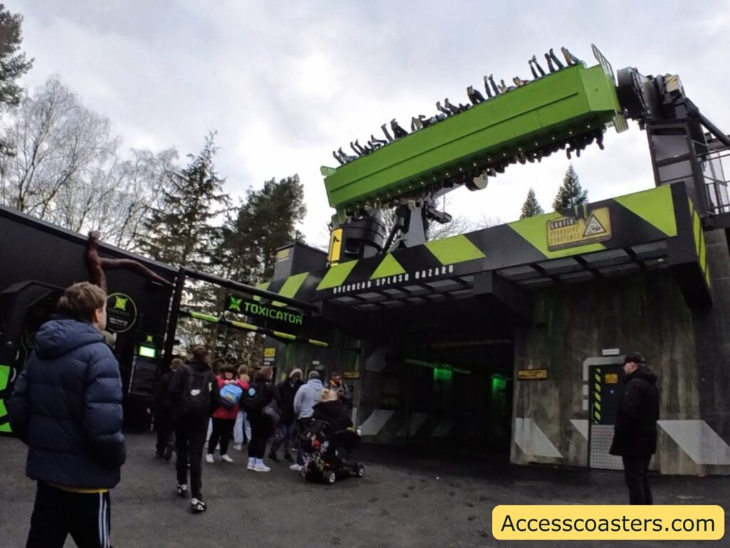 A group of people is gathered near an amusement park ride called "Toxicator." The ride features a large, green, rotating arm with seats attached, where riders' legs are visible sticking out as they are upside down. The entrance to the ride is designed with a toxic waste theme, featuring warning signs and hazard stripes. The surrounding area has trees and a cloudy sky. A person in a blue jacket and another in a black coat are visible among the crowd, along with a stroller. The website "Accesscoasters.com" is displayed in the bottom right corner.
