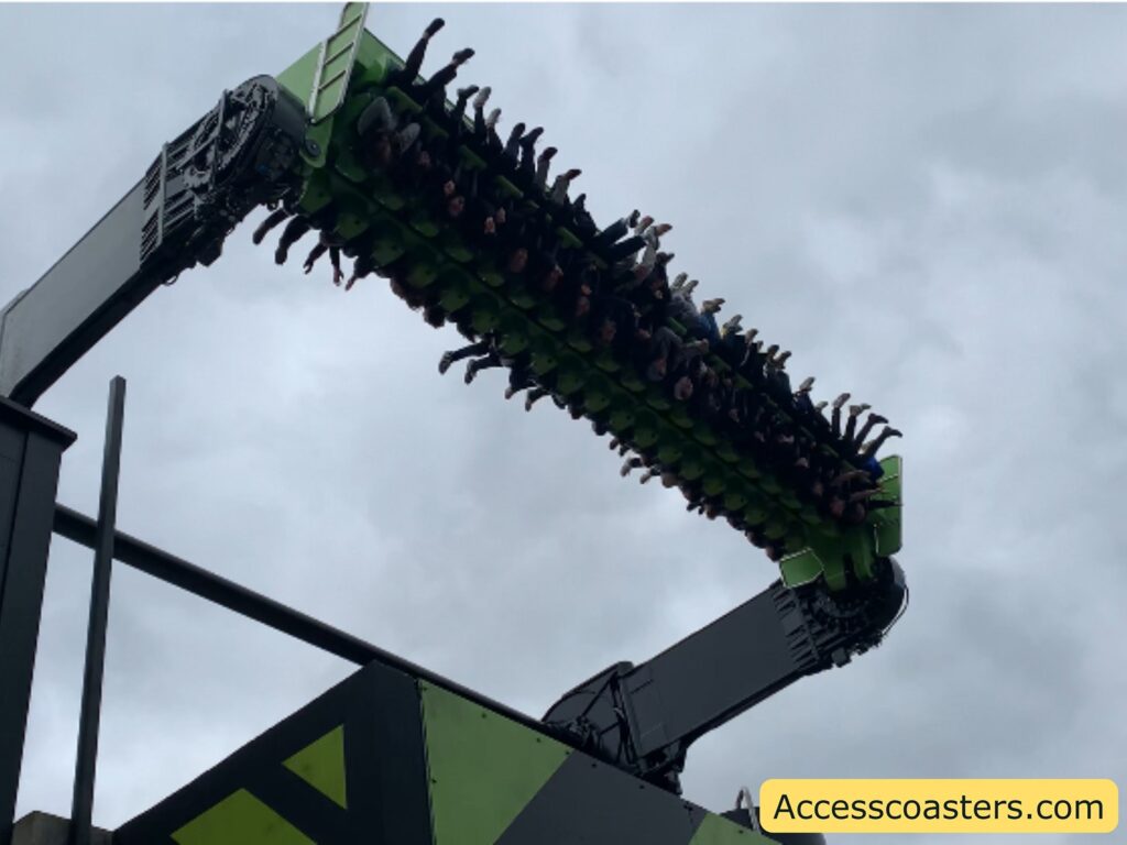 A group of people are on a thrill ride, suspended upside down in the air. The ride has a green and black color scheme, with two large mechanical arms holding the seating platform. The sky is overcast with gray clouds. In the bottom right corner, there is a yellow label with the text "Accesscoasters.com".