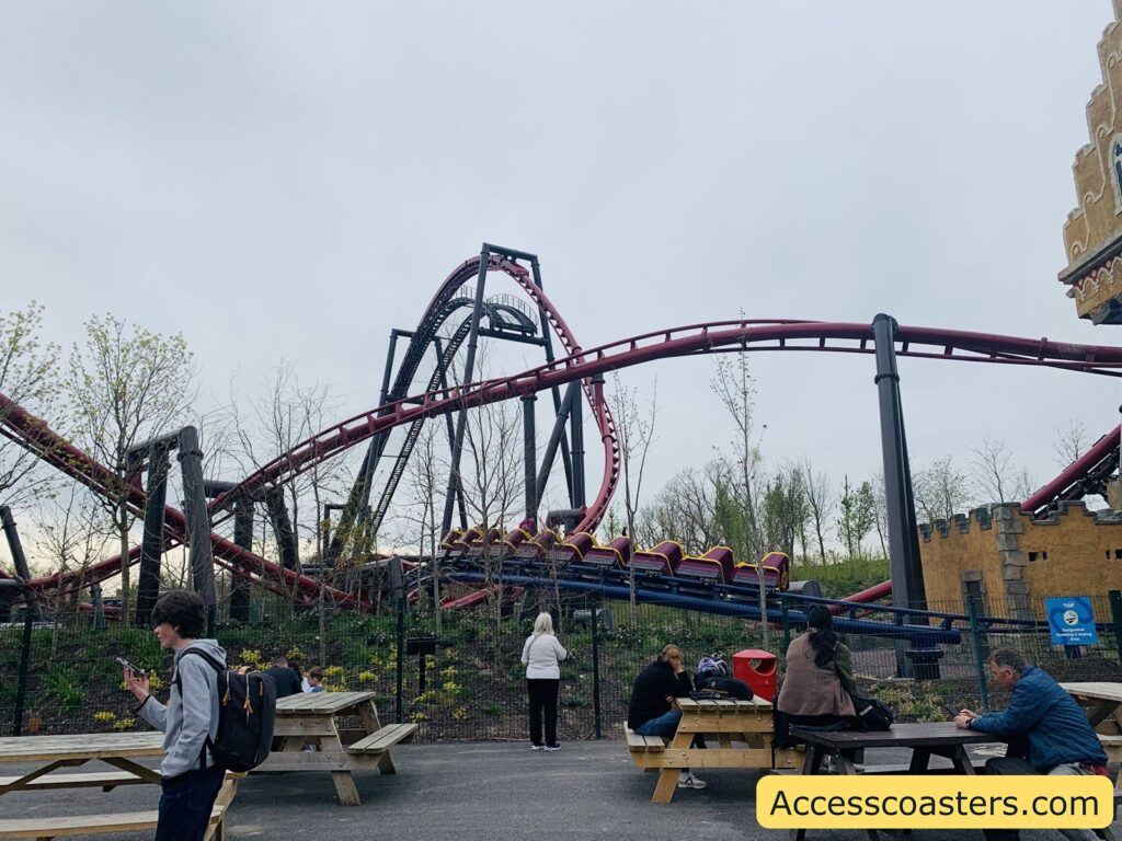 A roller coaster with red tracks and black supports, featuring a steep drop and several twists and turns. In the foreground, there are wooden picnic tables with people sitting and standing around them, some using their phones. The sky is overcast, and there are sparse trees and some castle-like structures on the right side.