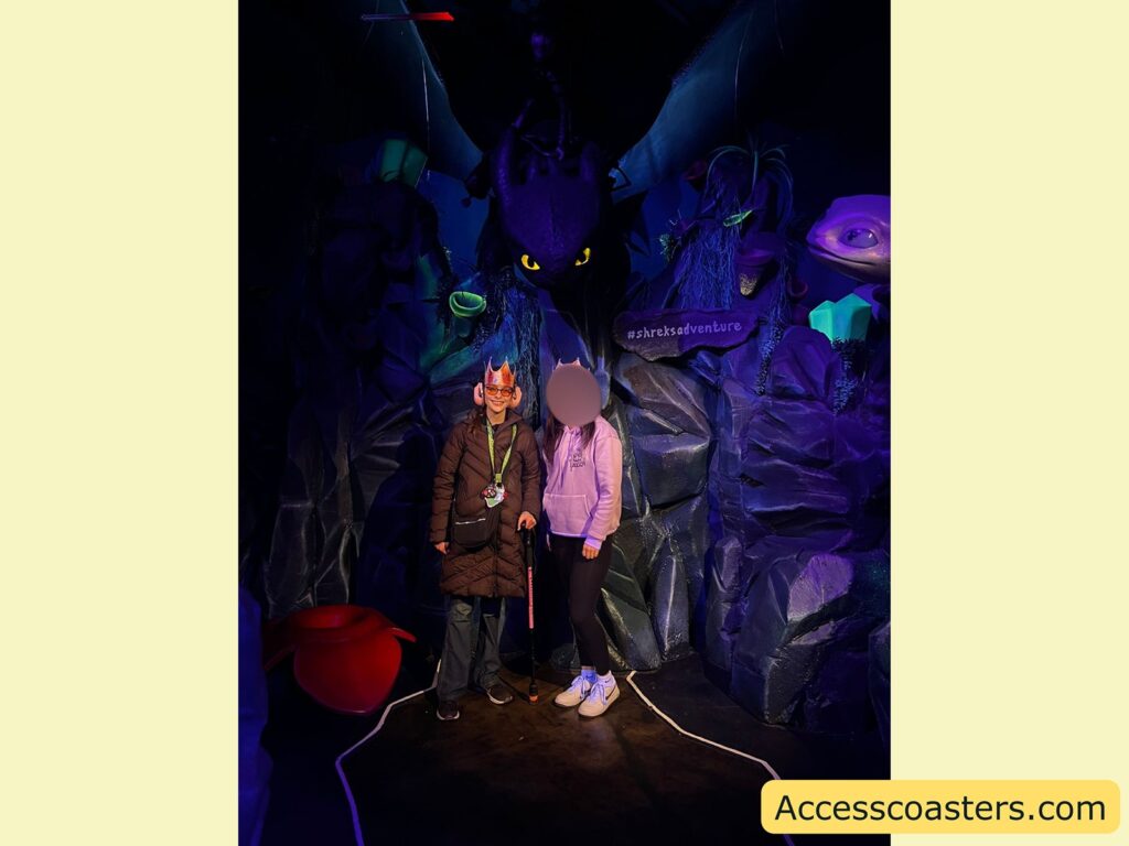 wo people standing in a themed, fantasy-like setting. Both are wearing paper crowns and smiling at the camera. The person on the left is wearing a long brown coat, glasses, and holding a pink cane, while the person on the right is in a light purple hoodie, black leggings, and white sneakers. Behind them, a large black dragon with glowing yellow eyes (resembling Toothless from "How to Train Your Dragon") looms overhead. The background is decorated with artificial rocks, glowing green crystals, and fantasy props.