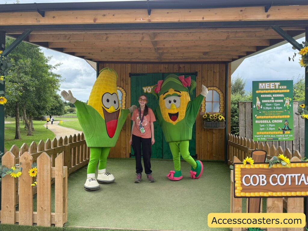 Outdoor photo of a person standing between two people in large corn-on-the-cob costumes, under a wooden shelter decorated with sunflowers. The corn characters have big cartoon eyes and smiling mouths; one has a pink bow and pink shoes, the other has a red tongue sticking out. The person in the middle is smiling, wearing glasses, a pinkish t-shirt, black pants, and hiking boots.

To the right, a green sign lists meet-and-greet times for "Kernel Kernel & Sweetie Corn" and "Russell Crow."