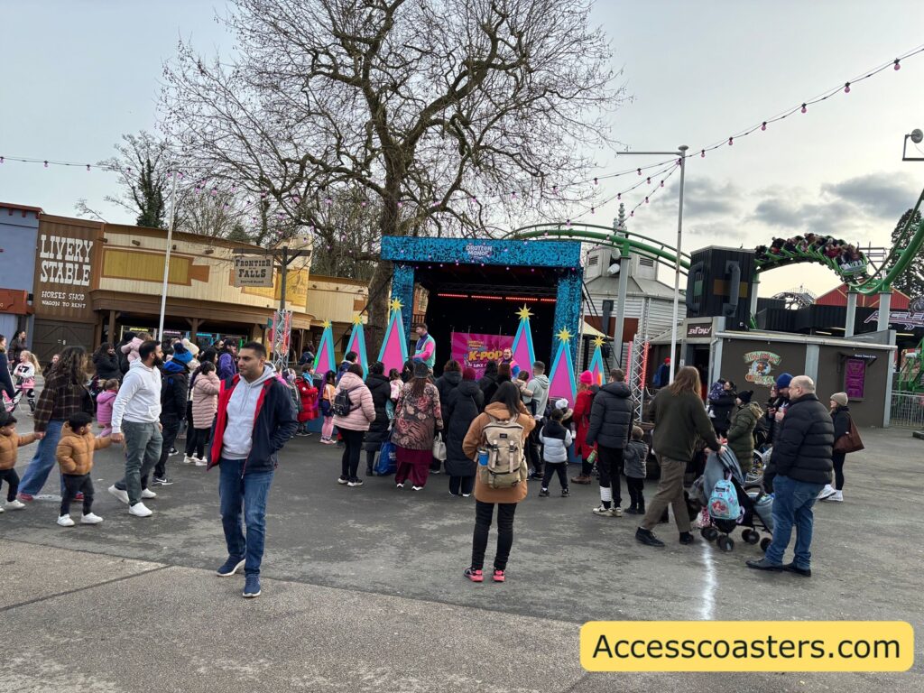 Crowd of adults and children gathered in an open paved area in front of a small outdoor stage decorated with bright, colorful panels, with string lights overhead and a roller coaster track visible in the background.