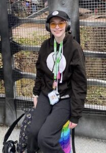 A picture of Iona white female, sat on a rainbow portable stool with a cap and headphones on smiling at the camera.