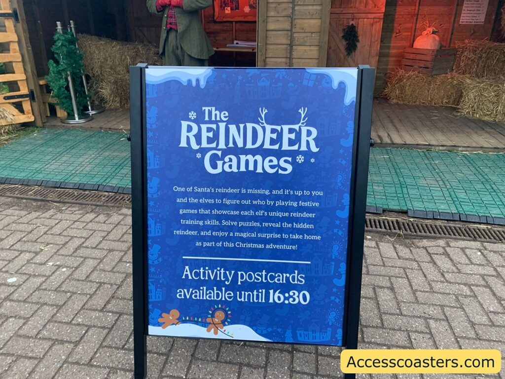 The image shows a signboard with a festive theme. The sign reads:
"The Reindeer Games
One of Santa's reindeer is missing, and it's up to you and the elves to figure out who by playing festive games that showcase each elf's unique reindeer training skills. Solve puzzles, reveal the hidden reindeer, and enjoy a magical surprise to take home as part of this Christmas adventure!
Activity postcards available until 16:30"
In the background, there is a wooden structure resembling a stable, with hay bales and a small Christmas tree. There is a person standing inside the structure, dressed in a festive outfit. The ground is paved with bricks, and there is a yellow label at the bottom right corner of the image that reads "Accesscoasters.com".