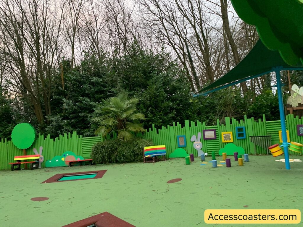 The image shows a children's play area with a colorful and playful design. The ground is covered with a soft, green surface, and there are various play elements scattered around. There are two benches with rainbow-colored slats, a series of small, colorful stepping posts, and a small trampoline embedded in the ground. The area is surrounded by a green fence with cartoonish decorations, including a rabbit figure and various shapes resembling trees and hills. In the background, there are tall trees and bushes. A large green canopy provides some shade over part of the play area. The bottom right corner of the image has the text "Accesscoasters.com".