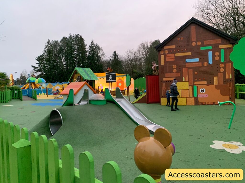 The image shows a colorful children's playground with various play structures. In the foreground, there is a small hill with two metal slides and a tunnel. The ground is covered with green and blue rubber safety flooring. To the right, there is a large wall painted with a whimsical design featuring a cow and geometric shapes. In the background, there are more play structures, including a house-like structure with a green roof and a pirate flag. There are a few people, including children and adults, walking around the playground. The area is surrounded by green fencing, and there are tall trees in the background. The sky is overcast. In the bottom right corner, there is a watermark that reads "Accesscoasters.com".