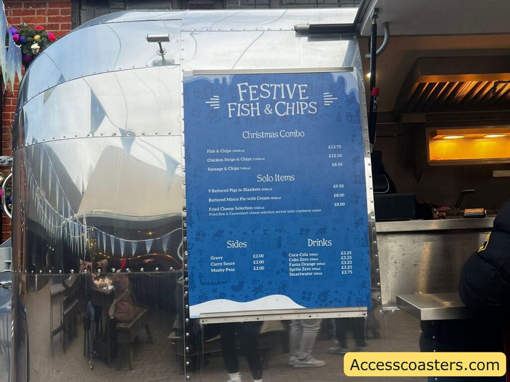 The image shows a food truck with a shiny metallic exterior. Attached to the side of the truck is a blue menu board titled "Festive Fish & Chips." The menu lists various food items and their prices.
Under "Christmas Combo," the options are:
- Fish & Chips for £13.75
- Chicken Strips & Chips for £12.50
- Sausage & Chips for £8.50
Under "Solo Items," the options are:
- 9 Battered Pigs in Blankets for £9.50
- Battered Mince Pie with Cream for £8.00
- Fried Cheese Selection for £8.00
Under "Sides," the options are:
- Gravy for £2.00
- Curry Sauce for £2.00
- Mushy Peas for £2.00
Under "Drinks," the options are:
- Coca-Cola 500ml for £3.25
- Coke Zero 500ml for £3.25
- Fanta Orange 500ml for £3.25
- Sprite Zero 500ml for £3.25
- Smart water 500ml for £2.75