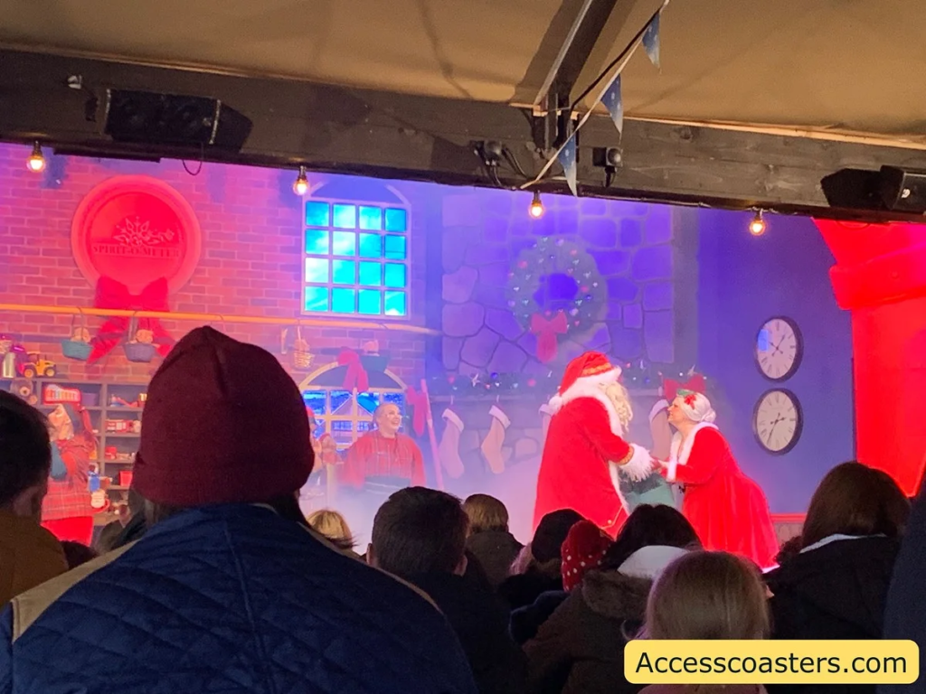 image shows a Christmas themed stage backdrop with Santa and mrs cluse holding hands. you can see heads of people in the audience watching the show