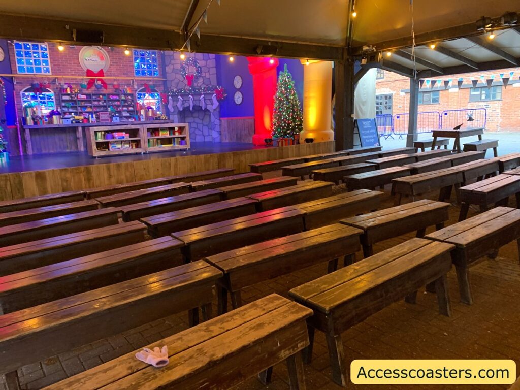 in the image you can see six rows of brown benches without backs. infant of the benches you can see the stage with a Christmas workshop scene set up.