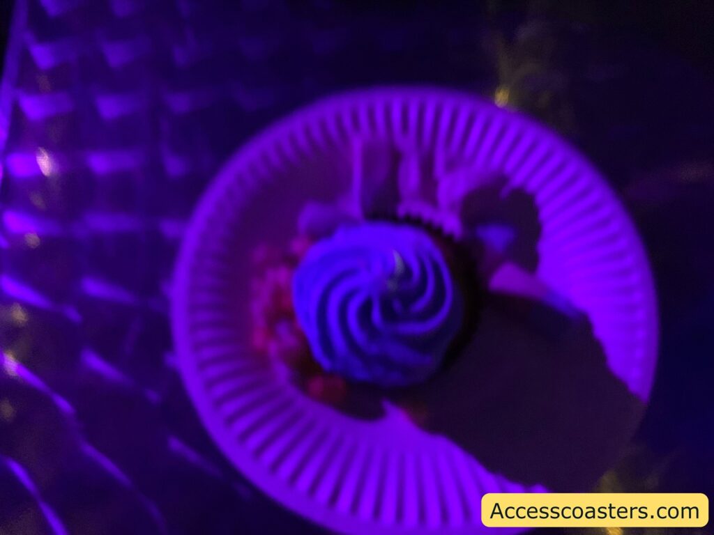 A paper plate with a cupcake on it, illuminated by purple lighting. The cupcake has a swirl of frosting on top. The background appears to be a textured surface, possibly a tablecloth or mat. In the bottom right corner, there is a yellow label with the text "Accesscoasters.com".