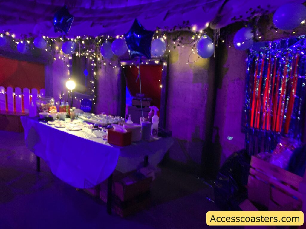A festive setup with a table covered in a white tablecloth, displaying various food items and drinks. The table is illuminated by a bright lamp, and there are decorations including balloons and string lights hanging above. The atmosphere is enhanced by a purple and blue lighting effect. On the right side, there are shiny, metallic streamers hanging on the wall. The bottom right corner of the image has a yellow label with the text "Accesscoasters.com".