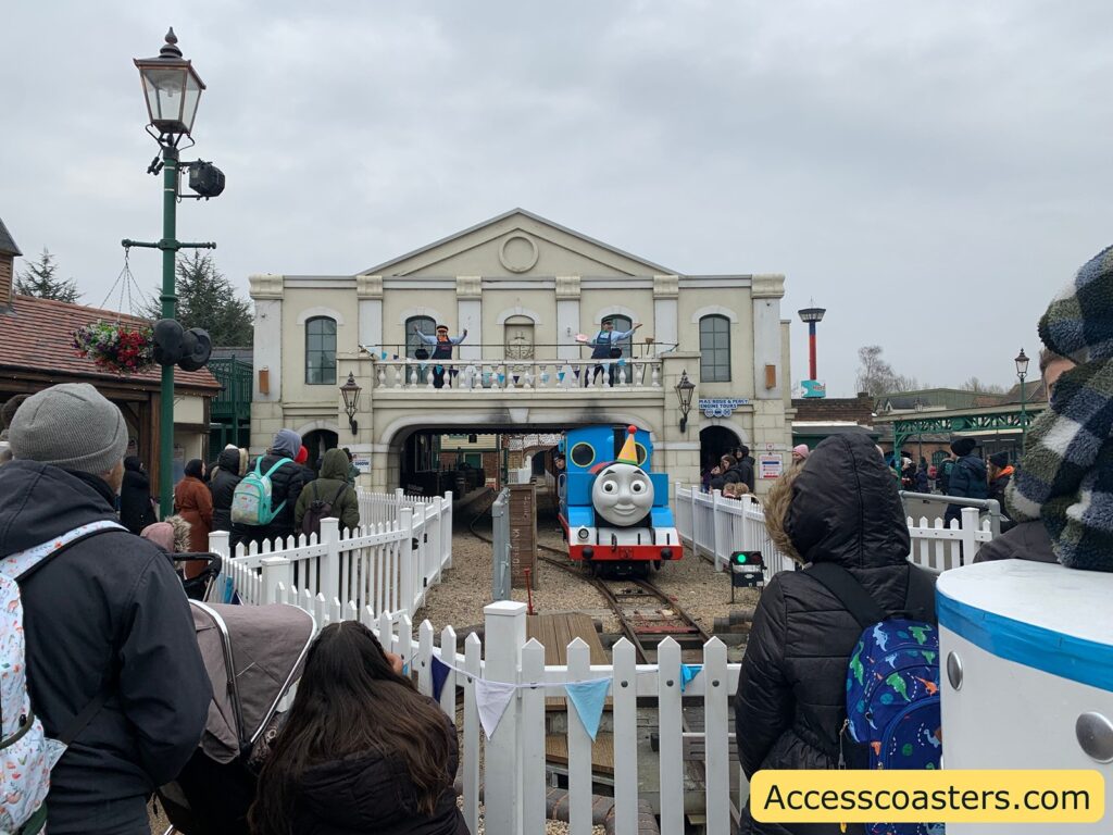 A crowd of people, dressed in winter clothing, is gathered around a small railway track. In the center, a life-sized model of Thomas the Tank Engine is positioned on the tracks, facing the crowd. Behind Thomas, there is a building designed to look like a train station with a balcony. On the balcony, a person dressed as a train conductor is waving to the crowd. The area is fenced with white picket fencing, and there are lampposts and a hanging flower basket nearby. The sky is overcast, suggesting a cold day. In the bottom right corner, there is a yellow banner with the text "Accesscoasters.com".