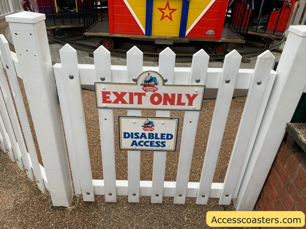 easy access entrance and exit for ladys carousel White picket fence with two signs attached. The top sign reads "EXIT ONLY" in red letters, and the bottom sign reads "DISABLED ACCESS" in blue letters. Both signs feature a small image of Thomas the Tank Engine with the text "Thomas Land" above it. Behind the fence, there is a colorful train ride with a red, yellow, and blue design featuring a star. The ground is covered with a textured surface. In the bottom right corner, there is a yellow rectangle with the text "Accesscoasters.com" in black.