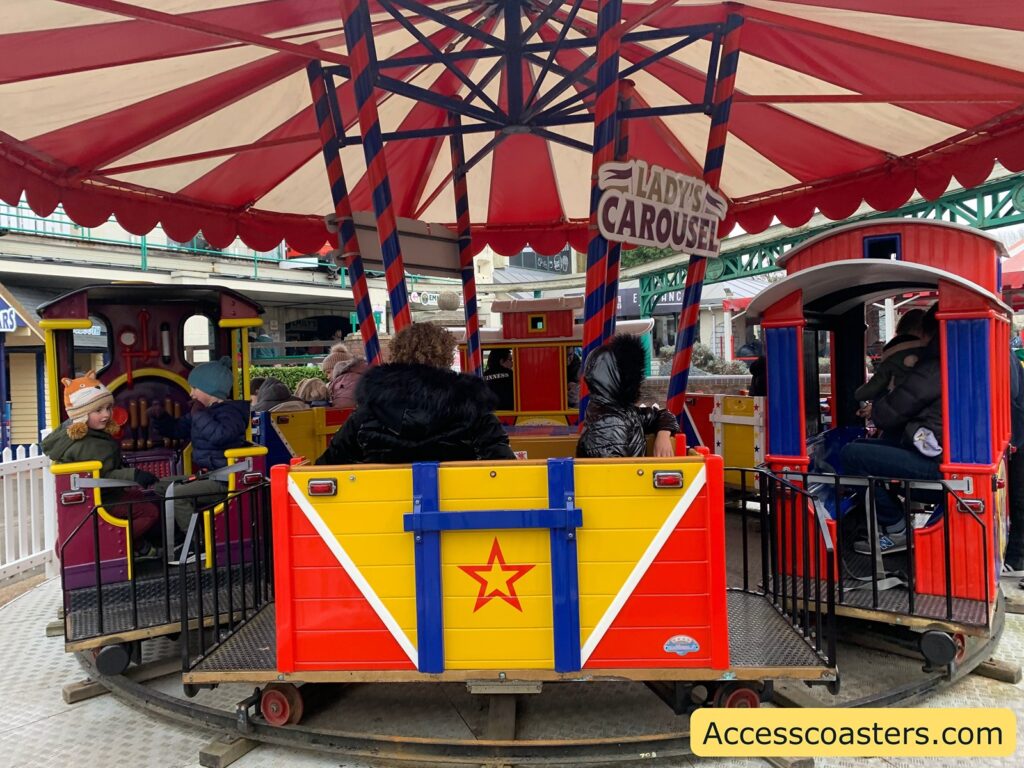 A colorful carousel ride with a circus theme. The carousel features small train-like cars with bright red, yellow, and blue colors. Each car has a star design on the side. There are several children and adults seated inside the cars, bundled up in winter clothing, indicating a cold day. The carousel is under a red and white striped canopy. A sign hanging from the canopy reads "Lady's Carousel." In the background, there are some buildings and a white picket fence surrounding the ride. In the bottom right corner, there is a yellow banner with the text "Accesscoasters.com."
