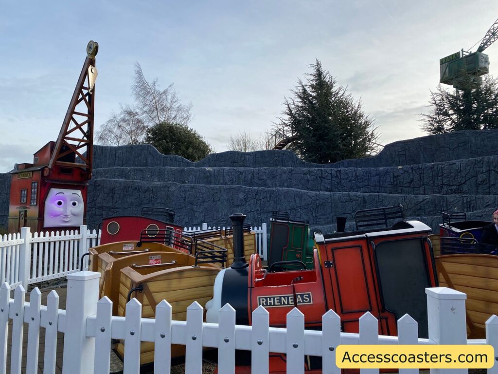 A small amusement park ride designed to look like a train with several carriages, each painted in different colors such as red, green, and brown. The train is named "RHENEAS," as indicated on the side of the red carriage. The ride is surrounded by a white picket fence. In the background, there is a large, animated face on a building resembling a crane, with a sign that reads "CRANKY" on the crane arm. The setting includes artificial rocky terrain and some trees. The sky is overcast. In the bottom right corner, there is a watermark with the text "Accesscoasters.com."