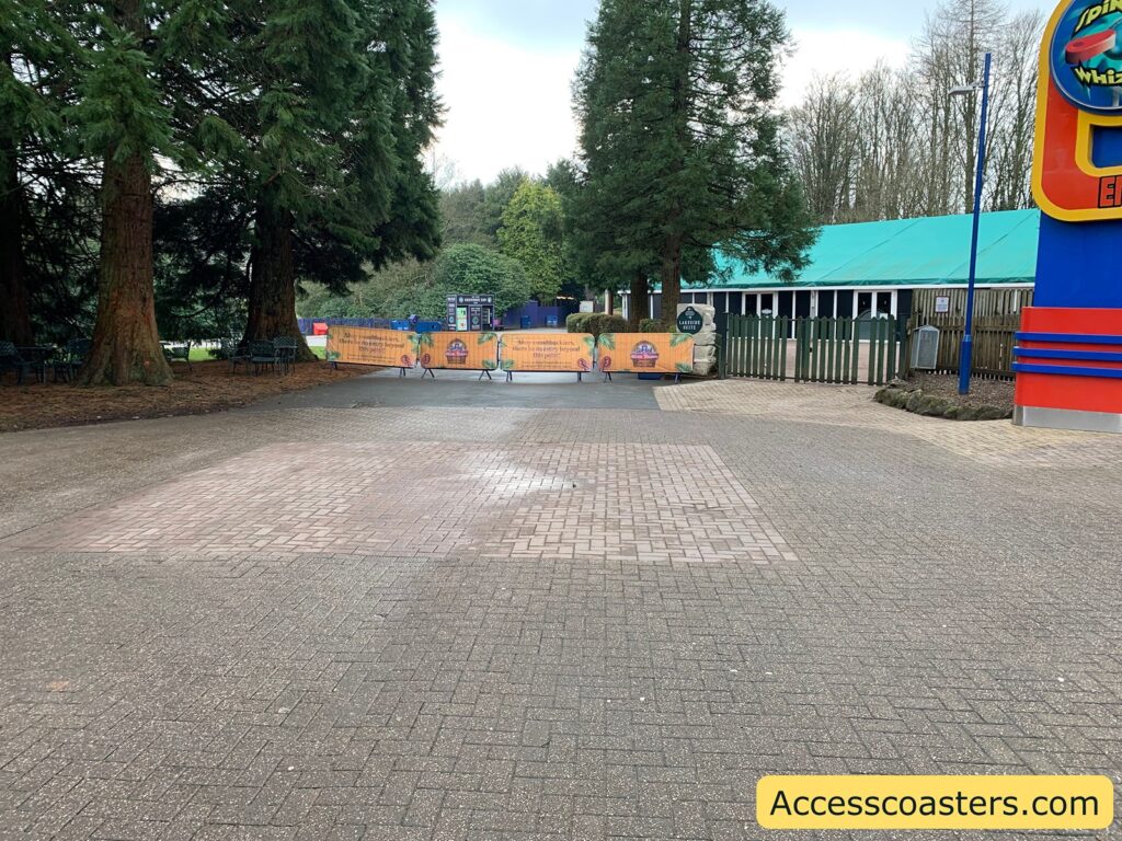 A paved pathway leads to a closed-off area with a temporary barrier featuring colorful signage. The barrier has images and text, indicating a closed off area. On the right, there is a building with a teal roof and a fence. Tall trees surround the area, and in the background, more trees and greenery are visible. In the bottom right corner, there is a yellow banner with the text "Accesscoasters.com".