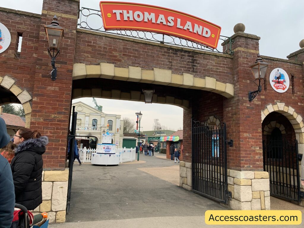 Entrance to Thomas Land, featuring a brick archway with a red sign displaying "THOMAS LAND" in white letters. The archway is flanked by two lanterns and has decorative stonework. Inside, there are people walking around, and a white picket fence surrounds a large, white, cake-shaped structure. A building with a classic facade is visible in the background. A small circular sign with an image of Thomas the Tank Engine and the words "THOMAS LAND" is on the right side of the archway. The bottom right corner has a yellow rectangle with the text "Accesscoasters.com".