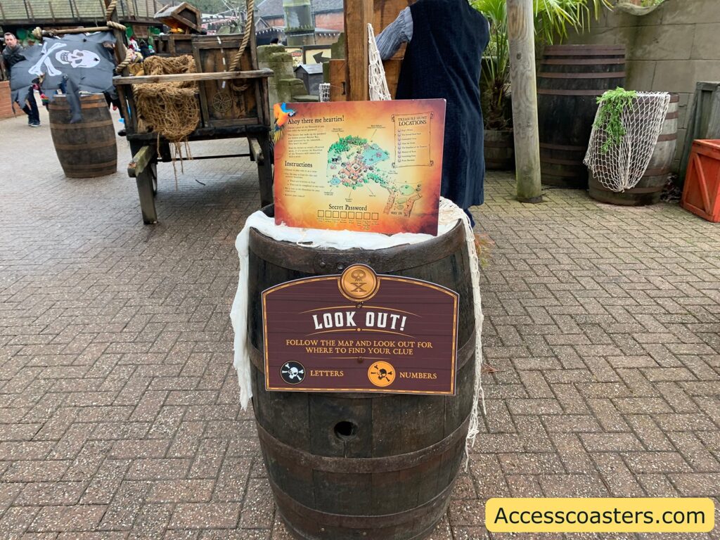 A wooden barrel is placed on a paved surface, with a sign attached to it that reads "LOOK OUT! FOLLOW THE MAP AND LOOK OUT FOR WHERE TO FIND YOUR CLUE" with the words "LETTERS" and "NUMBERS" below it, accompanied by a skull and crossbones symbol. On top of the barrel, there is a map with pirate-themed graphics and instructions. The background features more barrels, a cart with ropes and a pirate flag, and a person standing nearby. The website "Accesscoasters.com" is displayed in the bottom right corner.