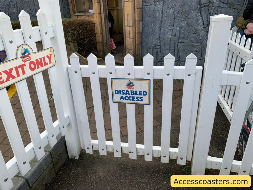 exit and easy access entrance for Blue Mountie Engines White picket fence with two signs. The sign on the left reads "EXIT ONLY" and features a small image of Thomas the Tank Engine. The sign on the gate in the center reads "DISABLED ACCESS" and also includes an image of Thomas the Tank Engine. The ground is paved with bricks, and there is a stone-textured wall in the background. A small portion of a person is visible behind the gate. The bottom right corner has a yellow rectangle with the text "Accesscoasters.com".
