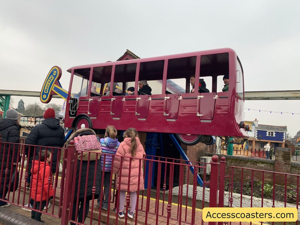 A group of people, including children and adults, are gathered around a red ride vehicle shaped like a bus. The bus is elevated on a track and has several passengers inside, visible through the windows. The ride is part of an amusement park setting, with colorful buildings and decorations in the background. The people outside the ride are dressed in winter clothing, suggesting a cold day. A sign on the ride reads "Bertie." The website "Accesscoasters.com" is displayed in the bottom right corner of the image.