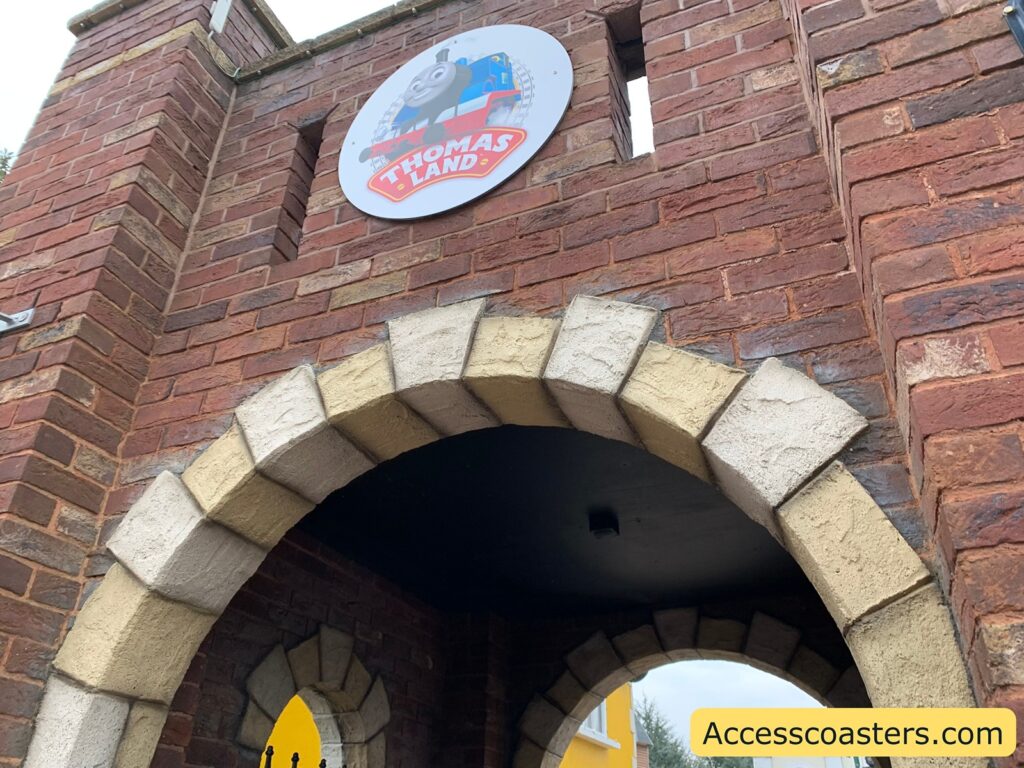 Brick archway with a circular sign above it featuring Thomas the Tank Engine and the text "THOMAS LAND." The archway is part of a larger brick structure with a small rectangular window above the sign. The bricks are a mix of red and brown tones, and the arch itself is outlined with lighter stone blocks. In the bottom right corner, there is a watermark with the text "Accesscoasters.com."