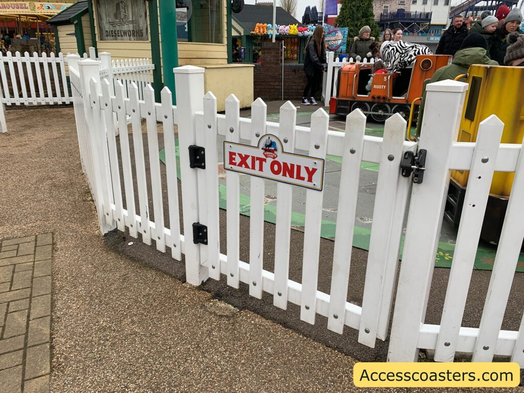 easy access entrance and exit White picket fence with a sign that reads "EXIT ONLY" in red letters. Behind the fence, there is a small amusement park ride featuring a train with the name "RUSTY" and the number "5" on it. Several people are visible near the ride, some wearing winter clothing. In the background, there is a building with a sign that says "DIESELWORKS." The ground is a mix of paved and textured surfaces. At the bottom right corner, there is a yellow banner with the text "Accesscoasters.com."