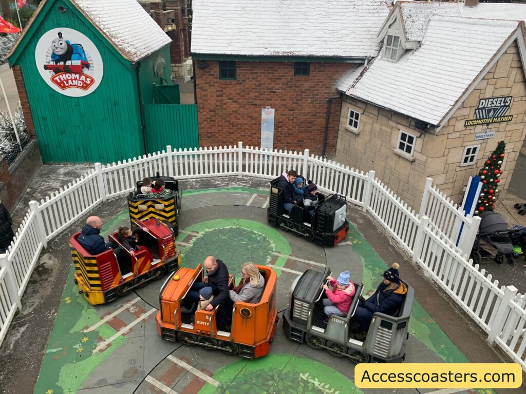 A small amusement ride featuring miniature train cars on a circular track. The ride is surrounded by a white picket fence. Each train car is uniquely colored and designed, with passengers seated inside. The area is themed around Thomas the Tank Engine, as indicated by the "Thomas Land" sign on a green building with a snowy roof. Another building with a stone facade has a sign reading "Diesel's Locomotive Mayhem." The ground is decorated with a track pattern and some leaf designs. A Christmas tree with red and gold ornaments is visible near the stone building. The website "Accesscoasters.com" is displayed in the bottom right corner.