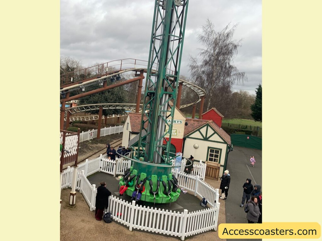 A small amusement park ride featuring a vertical drop tower with green seats arranged in a circular formation at the base of a tall, green metal structure. The ride is surrounded by a white picket fence. In the background, there is a small building with a sign that reads "Franky's Drop Tower" and a roller coaster track. Several people are standing around the ride, some with children, and a few are wearing winter clothing. The sky is overcast, and there are trees and a grassy area in the distance. The image has a yellow border with the text "Accesscoasters.com" at the bottom right corner.