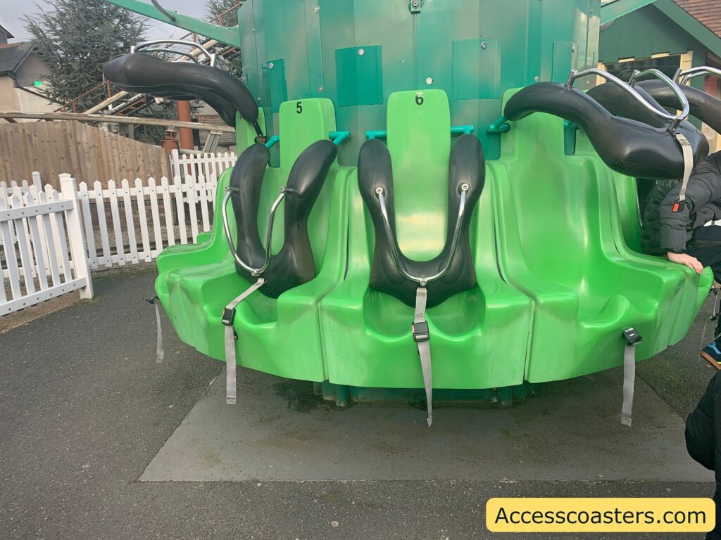 A row of bright green amusement park ride seats with black over-the-shoulder restraints and seat belts. Each seat is numbered, with visible numbers 5 and 6. The seats are part of a circular ride structure, likely a drop tower or similar attraction. In the background, there is a white picket fence and some trees. A person wearing a dark jacket is partially visible on the right side of the image. The bottom right corner has a yellow rectangle with the text "Accesscoasters.com".