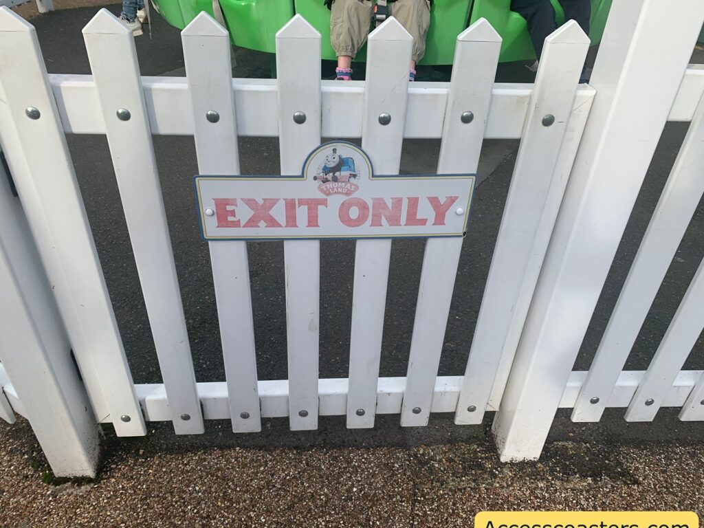 image of exi gate and easy access entrance for crankys drop tower. White picket fence with a sign that reads "EXIT ONLY" in red letters. Above the text, there is a small image of Thomas the Tank Engine with the words "THOMAS LAND" written below it. Behind the fence, there are people sitting on a green ride. The ground is paved with dark asphalt, and there is a small patch of gravel in the foreground.