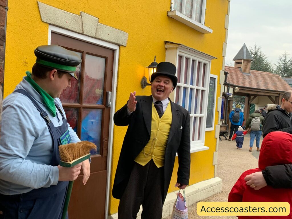 A lively scene of sir topham hat and rusty standing outside a bright yellow building. The man on the left is dressed as a worker, wearing a cap, a green bandana, and overalls, holding a broom. The man on the right is dressed in a formal outfit with a black top hat, a yellow waistcoat, and a black coat, smiling broadly and gesturing with his hand. In the background, people are walking, including a child in a red coat holding an adult's hand. The building has a wooden door and a window with colorful panes. The website "Accesscoasters.com" is written at the bottom right corner of the image.