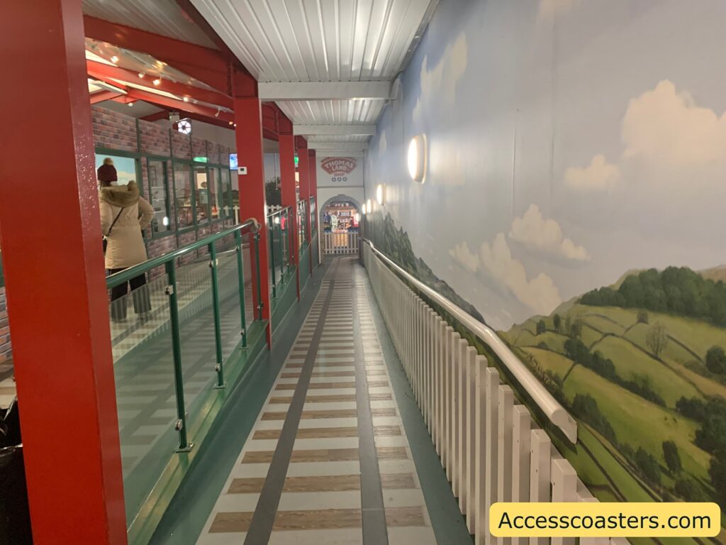 A long, indoor ramp with a gentle slope, featuring a handrail on the right side. The left side has a glass barrier with red metal posts. The wall on the right is painted with a mural depicting a scenic countryside landscape with green hills and a blue sky with clouds. The ceiling is white with evenly spaced lights. At the end of the ramp, there is an entrance to a shop with a sign that reads "Thomas Land Shop." A person wearing a beige coat and a red hat is walking down the ramp. In the bottom right corner, there is a watermark with the text "Accesscoasters.com."
