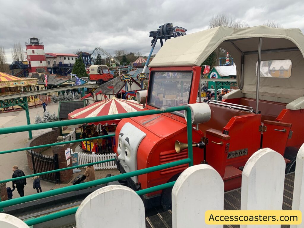 A theme park scene with a red, open-top vehicle resembling a character from the Thomas & Friends series, named Winston, as indicated by a nameplate on its side. The vehicle has a face on the front and a beige canopy. In the background, there are various amusement park rides, including a red and white striped carousel and a lighthouse structure. The sky is overcast, and there are a few people walking around the park. In the bottom right corner, there is a watermark with the text "Accesscoasters.com".
