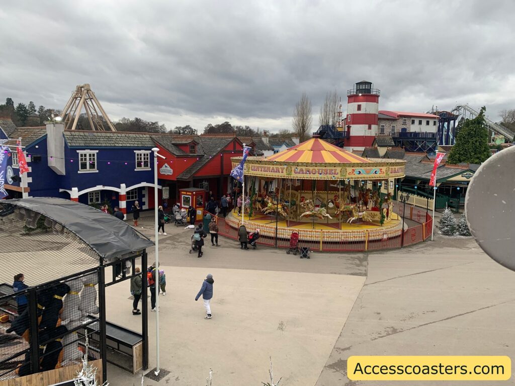 image of the middle of drayton manor with a vintage carousel in the center, featuring colorful horses and a yellow and red canopy. The carousel is surrounded by a red fence. In the background, there are various buildings, including a blue and red one with a sign that reads "Doughnut Factory." A lighthouse-like structure is visible, along with some roller coaster tracks. People are walking around the area, and a few are gathered near the carousel. The sky is overcast, and the ground is paved. In the foreground, there is a structure with a black mesh roof, possibly an enclosure or queue area. The bottom right corner has a yellow banner with the text "Accesscoasters.com."