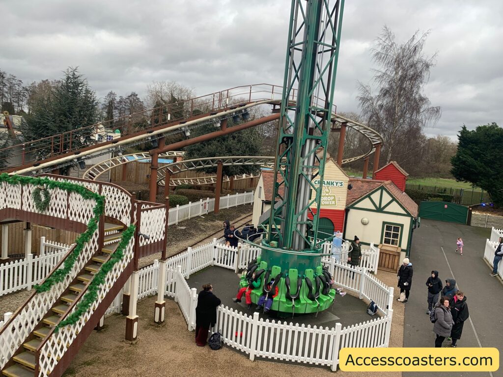 thomas land  with a drop tower ride named "Cranky's Drop Tower" surrounded by a white picket fence. The ride has green seats arranged in a circular formation at the base of a tall, green tower structure. Nearby, a roller coaster track with brown supports and white rails winds through the area. The surroundings include a small building with a red roof and a sign for the ride. People are walking around, dressed in winter clothing, indicating a cold day. The area is decorated with green garlands, and there are trees and cloudy skies in the background. A watermark in the bottom right corner reads "Accesscoasters.com".