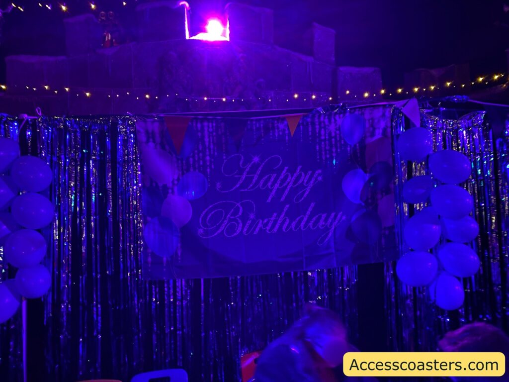 A festive scene with a "Happy Birthday" banner prominently displayed in the center. The area is decorated with shiny metallic streamers and clusters of balloons on either side of the banner. The lighting is dim with a strong purple hue, creating a party atmosphere. Above the banner, there are small string lights adding to the ambiance. In the bottom right corner, there is a watermark with the text "Accesscoasters.com".