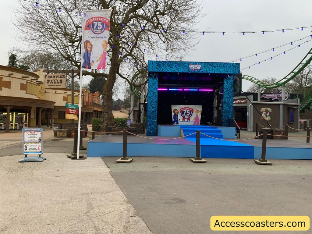 An outdoor stage area with a blue and black theme. The stage has a banner that reads "Celebrating 75 years of epic family fun" with cartoon characters on it. Above the stage, there are string lights with purple bulbs. To the left, there is a sign for "Frontier Falls" and a directional sign pointing to various attractions like "Haunting" and "Eating." In the background, there is a green roller coaster track. A signboard on the left provides entertainment information, and the ground is paved with a mix of concrete and asphalt. The image has a watermark in the bottom right corner that reads "Accesscoasters.com."