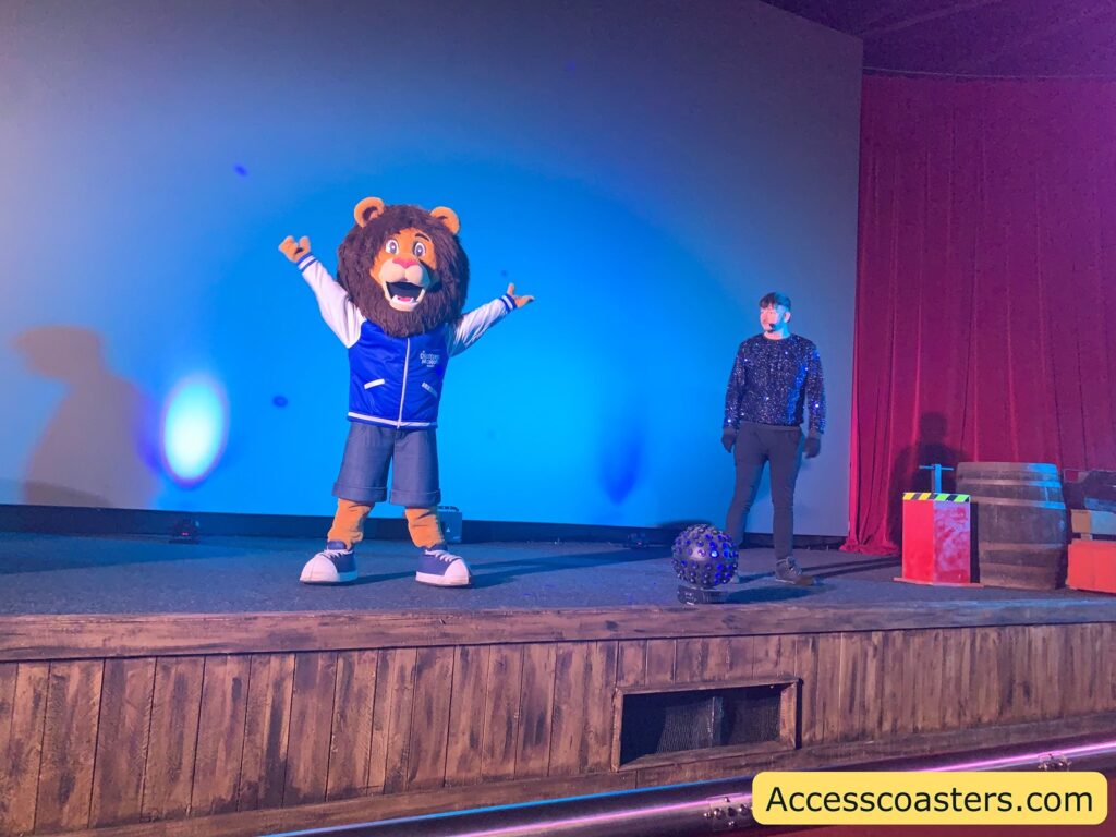 A stage with two performers. On the left, there is a Rory the lion, wearing a blue and white varsity jacket and denim shorts, with arms raised in an enthusiastic pose. On the right, a person dressed in a sparkly black top and black pants stands with a relaxed posture. The background is a plain blue-lit wall, and the stage is bordered by red curtains. There are some props on the stage, including a large wooden barrel and a red box with a yellow and black striped top. The website "Accesscoasters.com" is displayed in the bottom right corner.