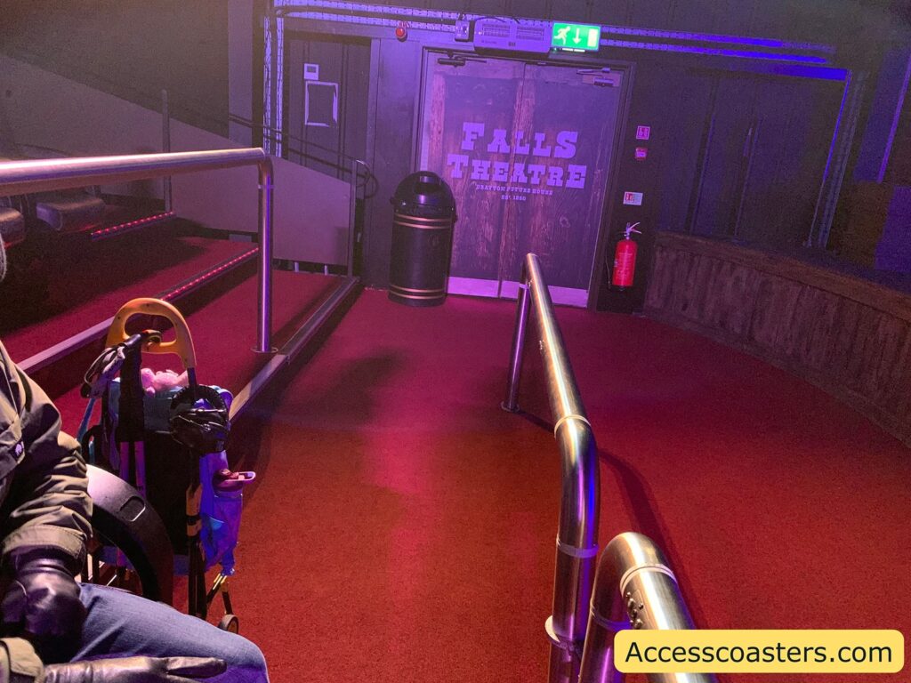A theater entrance with a door labeled "FALLS THEATRE" and "Phantom Falls Tours Est. 1926." The floor is carpeted in red, and there are metal handrails leading to the door. An emergency exit sign is above the door, and a fire extinguisher is mounted on the wall to the right. A trash can is positioned near the door. On the left side, part of a person in a wheelchair is visible, with a bag and some items attached to the wheelchair. The website "Accesscoasters.com" is displayed in the bottom right corner.