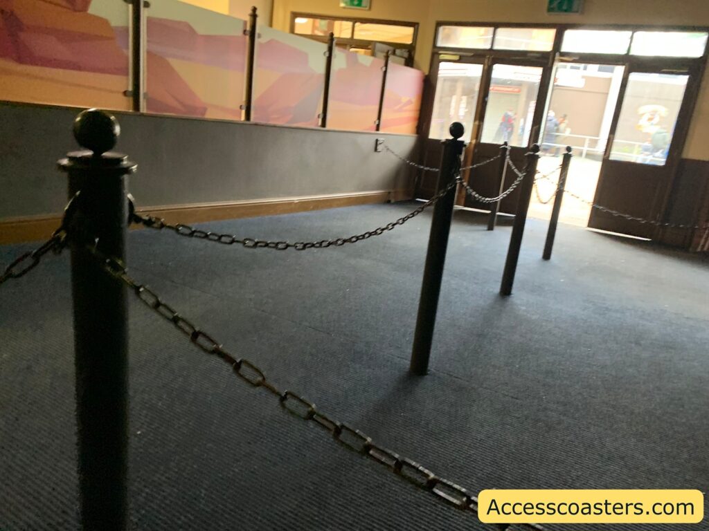 An indoor area with a dark carpeted floor and several black posts connected by chains, forming a queue line. The walls have a mural with abstract shapes in shades of pink, orange, and yellow. There are glass panels above the mural. At the far end, there are glass doors leading outside, where a few people can be seen in the background. In the bottom right corner, there is a yellow rectangle with the text "Accesscoasters.com".