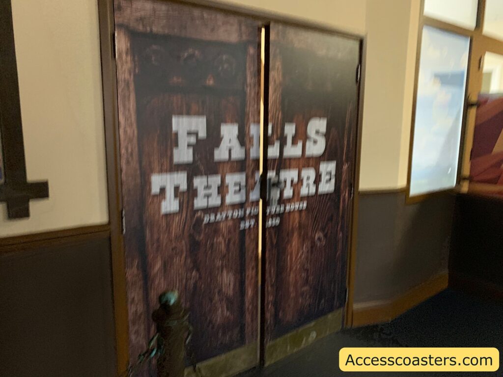 A set of double doors with a rustic wooden design. The text on the doors reads "FALLS THEATRE" in large white letters. Below that, in smaller text, it says "DAYTON FUN HOUSE" and "EST. 2020". To the right of the doors, there is a wall with a window showing a blue sky with clouds. In the bottom right corner of the image, there is a yellow rectangle with the text "Accesscoasters.com".