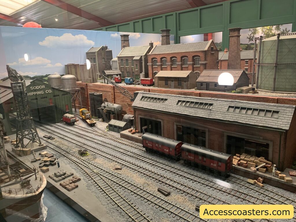 A detailed model railway scene depicting an industrial area. There are multiple railway tracks with two red mail train cars labeled "Sodor Mail" on one of the tracks. The setting includes several brick buildings, one of which has a sign that reads "Sodor Shipping Co." There are various industrial elements such as large storage tanks, a crane, and stacks of crates and sacks. The backdrop features a painted sky with clouds, and the scene is enclosed in a display case with a reflective surface. The website "Accesscoasters.com" is visible in the bottom right corner.