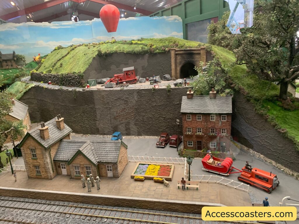 A detailed miniature model scene depicting a quaint village setting. In the foreground, there are two small stone buildings with slate roofs, resembling a train station or village houses. A group of four miniature figures stands on the platform, and a dog is nearby. A colorful flower bed is also present on the platform.

In the middle ground, a red sleigh is attached to an orange vehicle, possibly a snowplow or tractor. Two vintage cars are parked next to a three-story brick building with flower boxes under the windows.

In the background, a grassy hill with a tunnel entrance is visible. A red train car is positioned on the track leading into the tunnel. A hot air balloon hovers above the hill, and a small airplane is seen on the left side. The scene is enclosed within a display case, and the website "Accesscoasters.com" is printed in the bottom right corner.