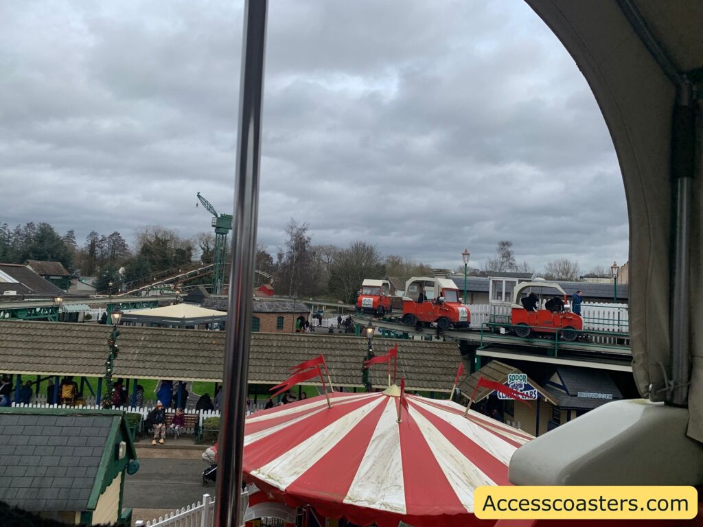 Overcast sky with gray clouds. In the foreground, a red and white striped tent top is visible. Behind it, there is a small elevated track with several red, vintage-style vehicles that resemble old-fashioned cars, each with a white canopy. People are riding in these vehicles. Below the track, there are buildings with green roofs and a sign that reads "Sodor Classic Cars." In the background, there are trees and a crane-like structure. The image has a watermark in the bottom right corner that reads "Accesscoasters.com."