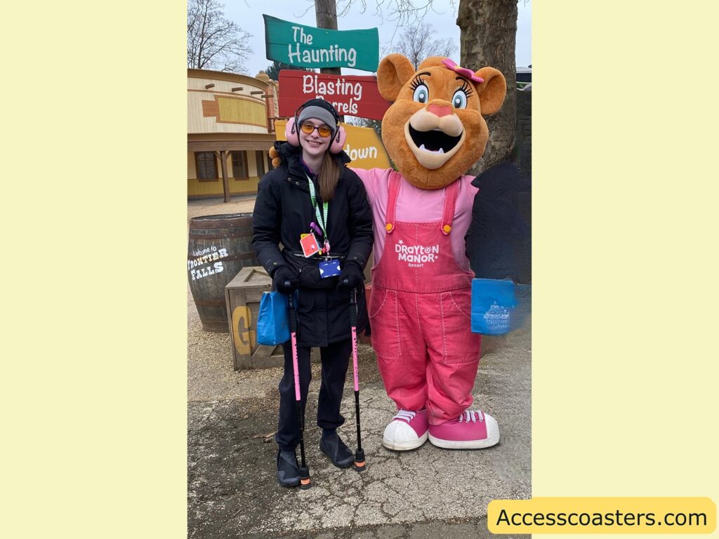 A person is standing next to Lory a large mascot character. The person is wearing a black coat, black gloves, and a black hat with earmuffs, along with orange-tinted glasses. They have a lanyard with several cards hanging from their neck and are holding a blue bag. The mascot is a bear wearing pink overalls with "Drayton Manor Resort" written on them, a pink shirt, and a pink bow on its head. In the background, there are signs that read "The Haunting," "Blasting Barrels," and "Sheriff's Showdown." A wooden barrel with "Welcome to Frontier Falls" written on it is also visible. The bottom right corner of the image has the text "Accesscoasters.com."