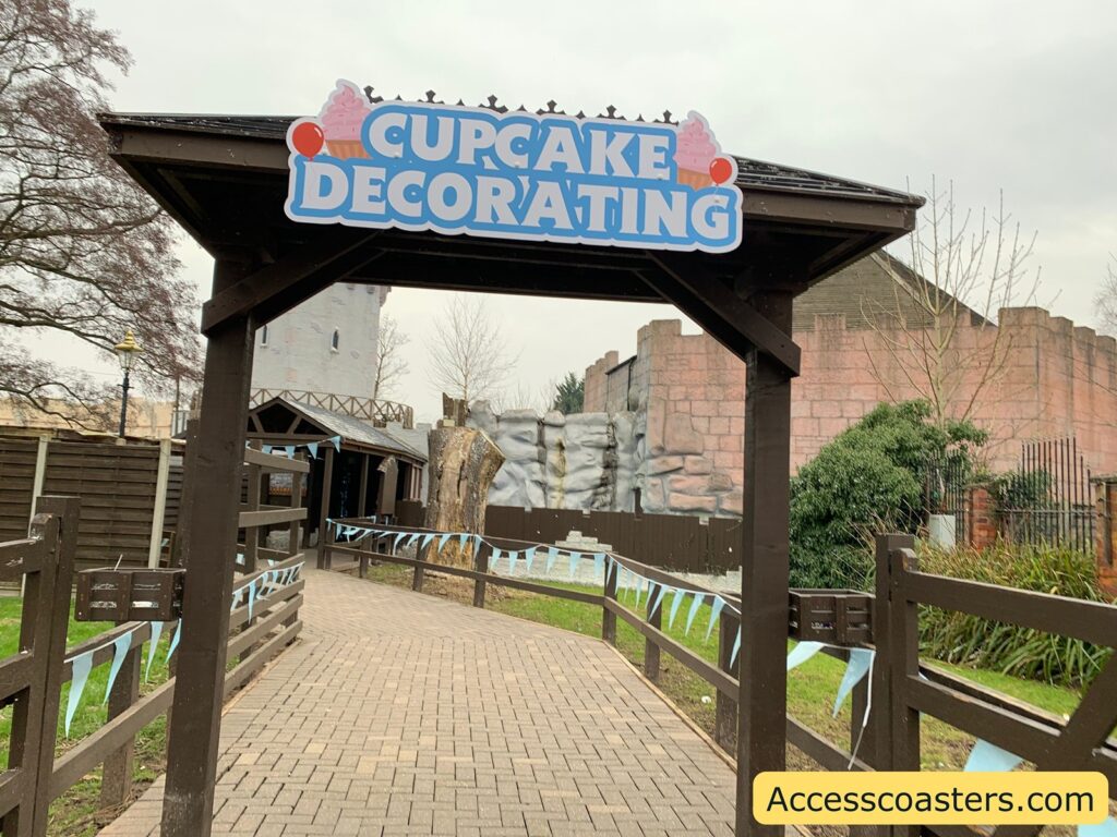 A wooden archway with a sign on top that reads "CUPCAKE DECORATING" in large blue letters, accompanied by images of a cupcake and an ice cream cone. The path beneath the archway is paved with bricks and is lined with wooden fences adorned with light blue triangular flags. In the background, there is a building with a castle-like facade, featuring stone textures and battlements. To the right, there is greenery, including bushes and trees. In the bottom right corner, there is a yellow rectangle with the text "Accesscoasters.com".
